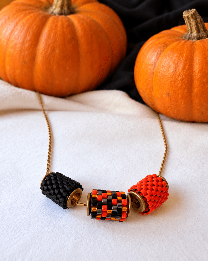 Halloween necklace with three beaded charms in black, orange and a mixed pattern with sparkling accents, displayed on white fabric with orange pumpkins and black fabric in the background – Fleur du Jour