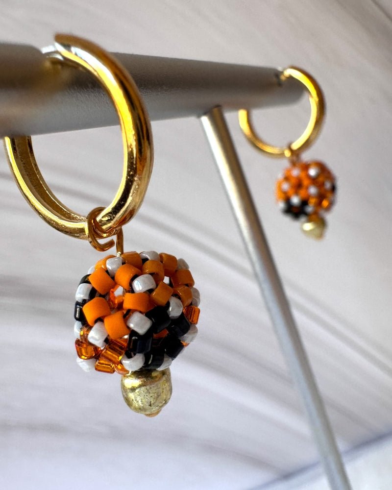 Gold-plated Halloween hoop earrings with spherical Miyuki beaded charms in orange, black and white with sparkling orange accents, displayed on a jewellery stand – Fleur du Jour